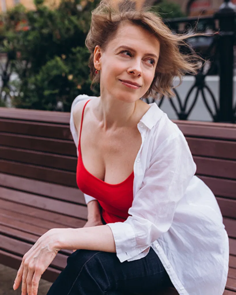 Anastasia dehors, assise sur un banc, portant un top rouge et une chemise bleue