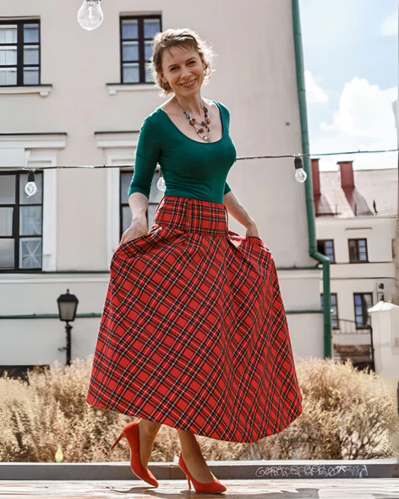 Anastasia souriante devant un bâtiment, portant un top vert et une longue jupe rouge à carreaux